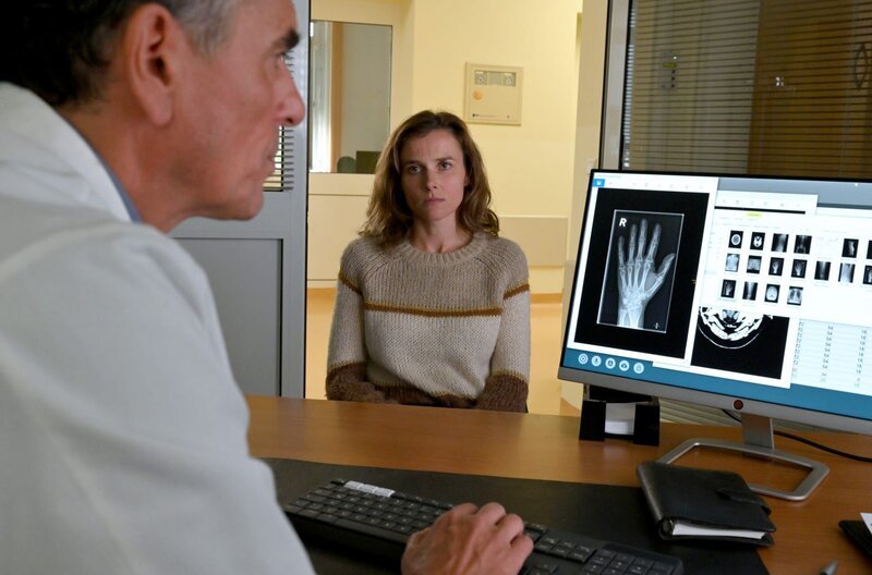 Karin Gorniak (Karin Hanczewski) ist beim Amtsarzt Dr. Herbst (Ahmad Mesgarha) um eine Ursache für ihre Schmerzen zu finden. – Bild: MDR/​MadeFor/​Hardy Spitz