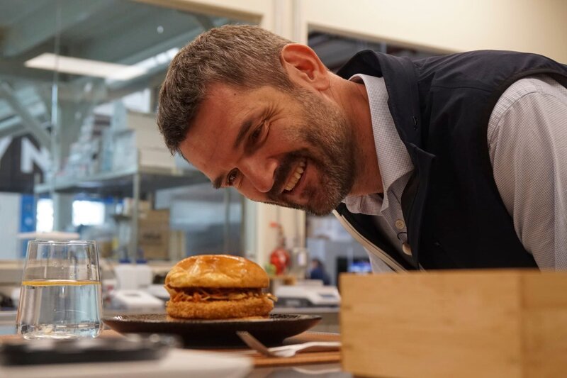 Dr. Mario Herger kostet einen veganen Chickenburger – die Rezeptur wurde entwickelt von einer künstlichen Intelligenz der Firma „NotCo“. – Bild: ServusTV