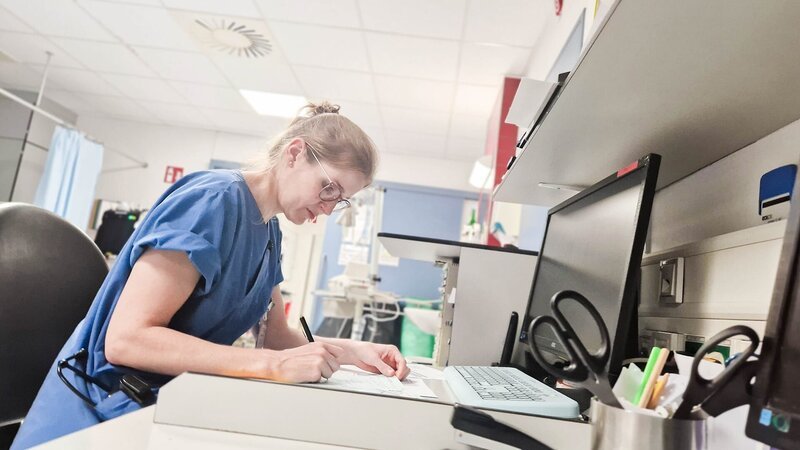 E7 – Female physician with blue scrubs writing a report while sitting at a desk with a computer in front of her. To the right there are scissors in a cup – Bild: Warner Bros. Discovery, Inc.
