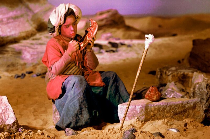 Im Orient lebt ein kleiner aufgeweckter Junge namens Muck, der einen Buckel hat und den alle verhöhnen und auslachen. In all seinem Jammer läuft er in die Wüste, um den Kaufmann zu suchen, der das Glück zu verkaufen hat. Den Kaufmann findet er nicht, aber dafür ein Zauberstöckchen und Zauberpantoffeln, mit denen er schnell wie der Blitz laufen kann. Märchenklassiker DEFA 1953 – Der kleine Muck (Thomas Schmidt) begutachtet die rasenden Zauberpantoffel und den verzauberten Stab, der Schätze findet. Beides hat er einer wundersamen Zauberin entwendet. – Bild: rbb/​Progress Film-Verleih/​Eduard Neu