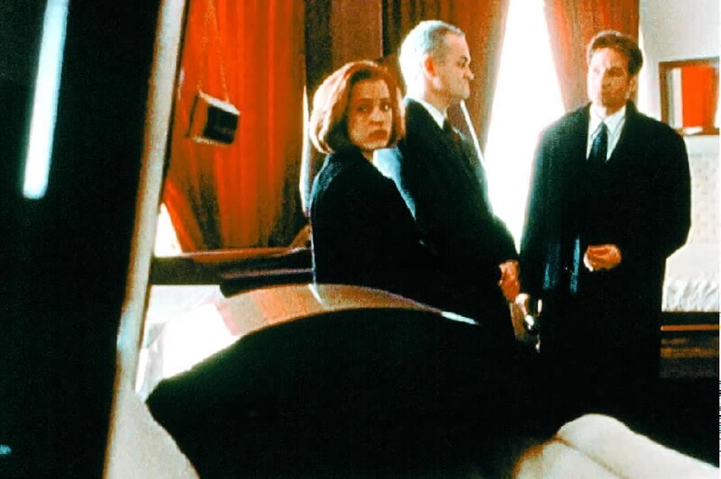 Mulder (David Duchovny, r.) und Scully (Gillian Anderson, l.) sind erstaunt, in einem kleinen Ort mit knapp 400 Einwohnern das größte Sarglager von ganz Texas vorzufinden. Der Inhaber (Forbes Angus, M.) führt die beiden hingegen stolz durch seinen Laden. – Bild: SYFY