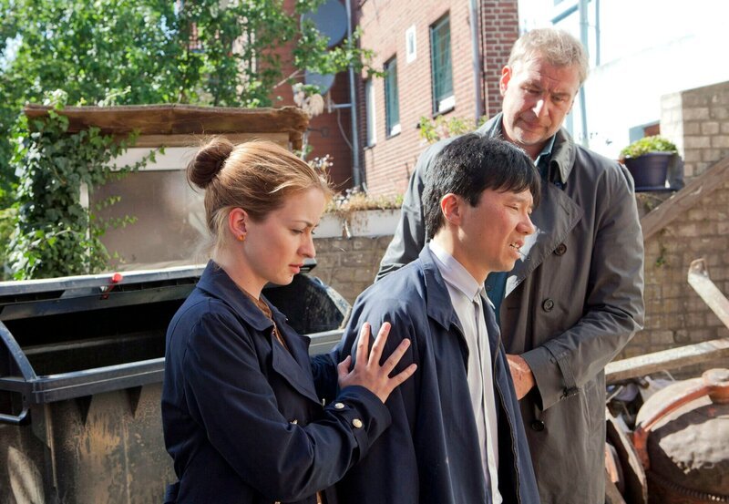 Nora (Loretta Stern, l.) und Jon (Frank Vockroth, r.) stützen Hu (Yuki Iwamoto, M.), den verzweifelten Vater des Ermordeten. – Bild: NDR/​ARD/​Georges Pauly