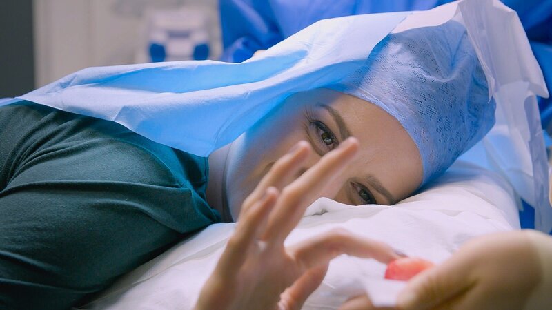 Close up shot of Charnelle M lying on treatment bed, touching the cyst that has just been removed from her neck. – Bild: Warner Bros. Discovery, Inc.