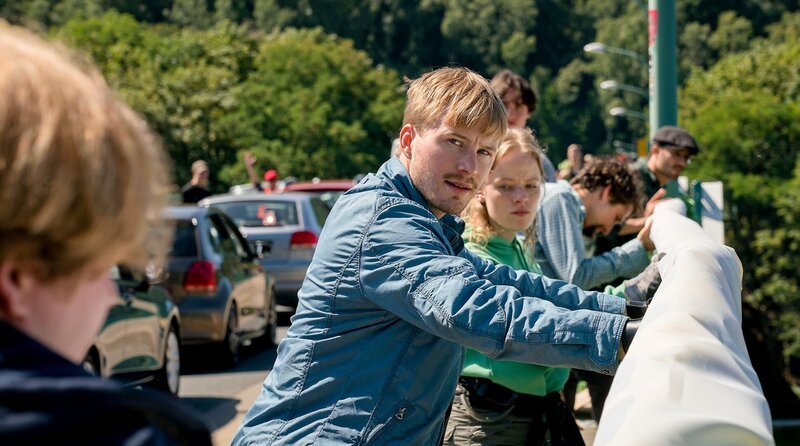Kevin Holzer (Tillmann Eckard, M.), entrollt zusammen mit Leonie Kaiser (Luna Kuse, h.) und weiteren Aktivist:innen (Komparsen) ein Transparent. – Bild: ARD/​Rudolf Wernicke
