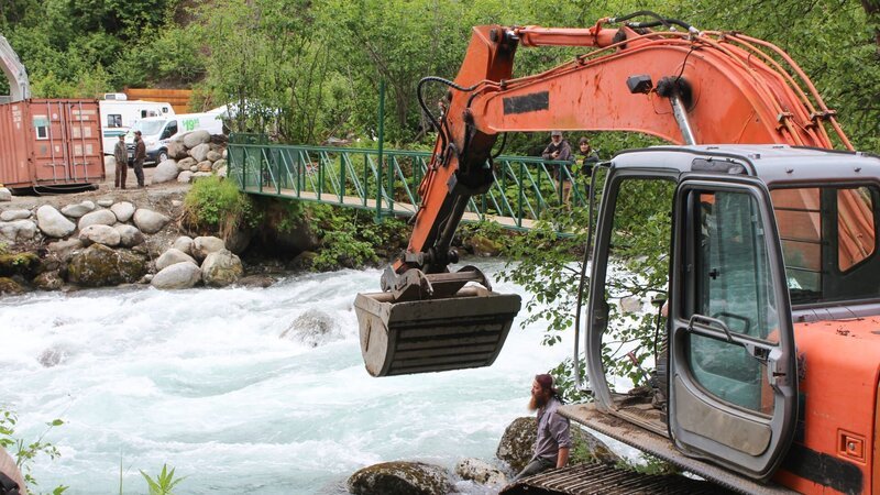 Weite Aufnahme von Bagger und Transportkiste, getrennt durch einen Fluss. – Bild: Scott Sandman /​ Discovery Channel /​ Discovery Communications, LLC