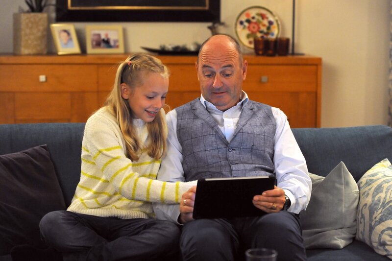 Franzi (Mia Löffler, KLD) sitzt mit ihrem Vater Hubert (Bernhard Ulrich) auf der Couch und zeigt ihm ein Spiel auf dem Tablet. – Bild: Marco Orlando Pichler /​ BR