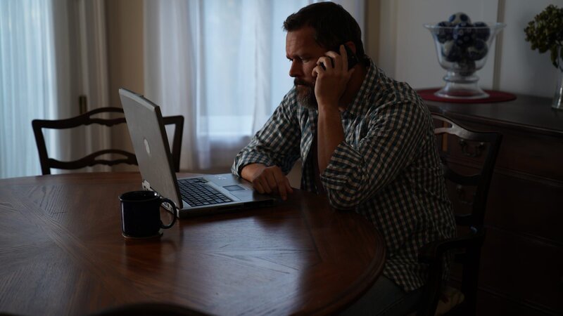 Recre of Michael sitting at dining table looking at computer on the phone with hand closest to camera. Side angle shot. – Bild: TLC