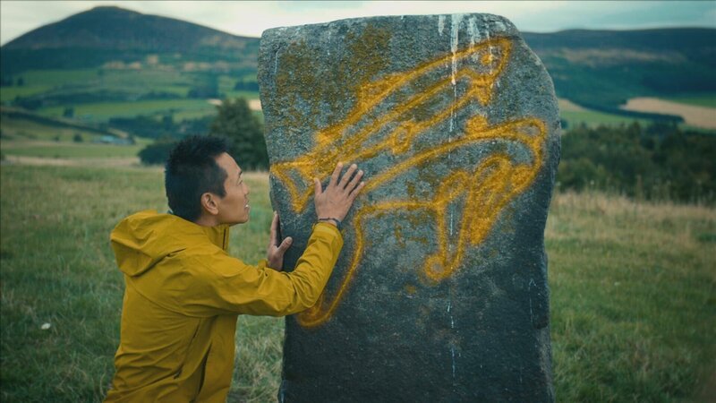 Albert Lin untersucht einen großen stehenden Felsen mit Einritzungen piktischer Symbole, die in diesem computergenerierten Bild grafisch verbessert und hervorgehoben wurden. Tap O’Noth ist im Hintergrund zu sehen. – Bild: National Geographic for Disney /​ Disney