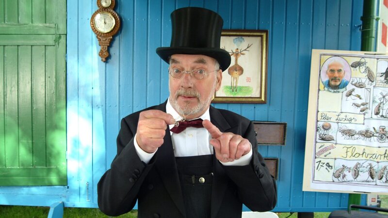 Peter (Peter Lustig), der neue Flohzirkusdirektor, zeigt eine kleine Kutsche, die gleich von einem Floh gezogen wird. – Bild: ZDF und Christiane Pausch