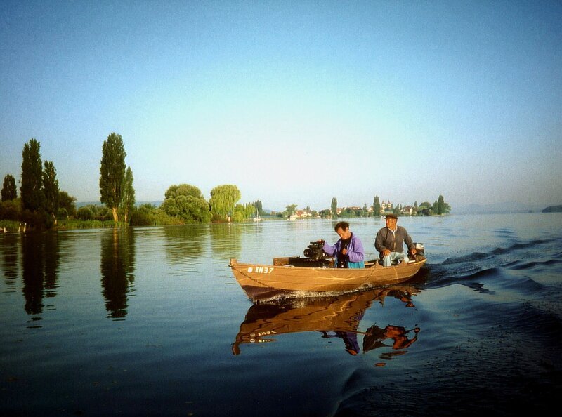 SWR-Kameramann im Boot eines Bodenseefischers. Im Hintergrund die Insel Reichenau. – Bild: BR/​SWR