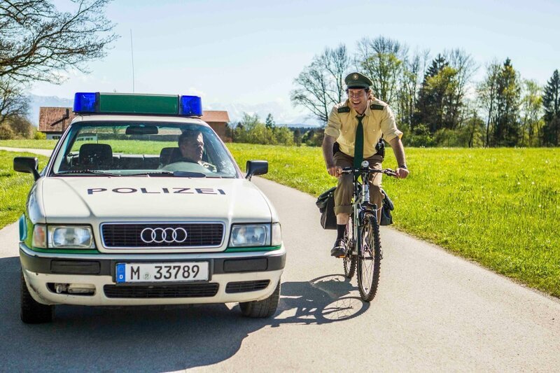 v.li.: Während Hubert (Christian Tramitz) im Dienstwagen fährt, muss Staller (Helmfried von Lüttichau) mit dem Fahrrad fahren. – Bild: TMG /​ Chris Hirschhäuser
