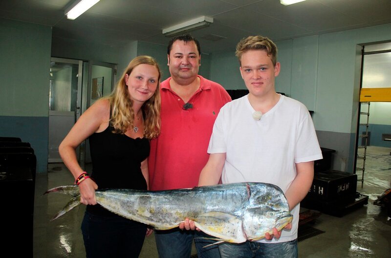 Küchenpraktikantin Elisabeth, Küchenchef Fritz und Küchenpraktikant Andreas posieren auf einem Markt in St. Vincent in der Karibik mit einem großen Fisch. – Bild: BR/​Gerrit Mannes