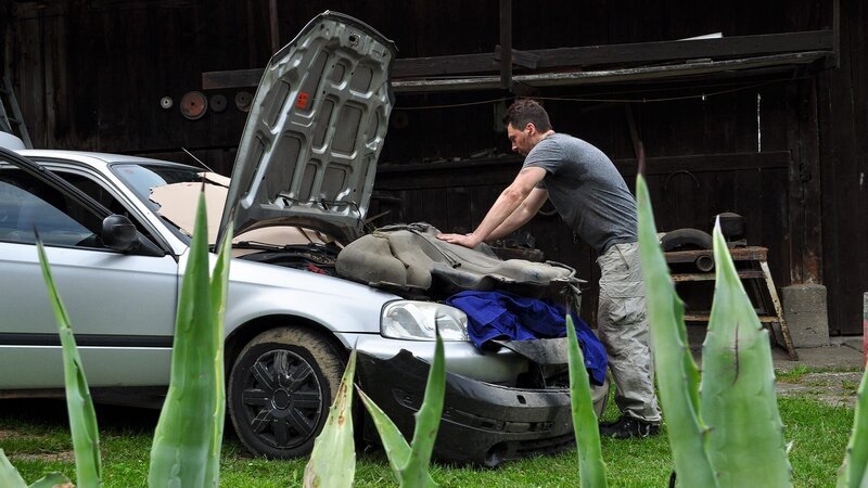 Ein Mann vor seinem Schuppen mit einem Auto mit offener Motorhaube und Werkzeug auf dem Tisch. – Bild: Warner Bros. Discovery