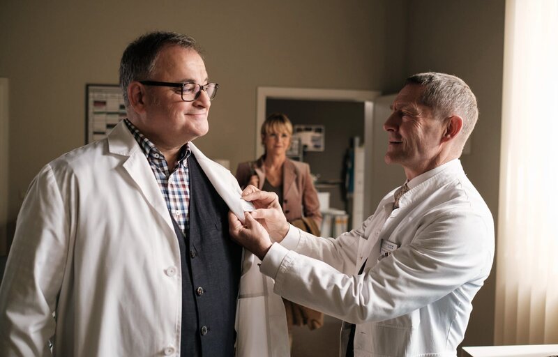 Hans-Peter Brenners (Michael Trischan, li.) ehemalige Prüferin Dr. Sylvia Jessel (Esther Esche, mi.) betritt gerade das Ärztezimmer, als Dr. Rolf Kaminski (Udo Schenk, re.) dem frischgebackenen Doktor das neue Namensschild anheftet. – Bild: MDR