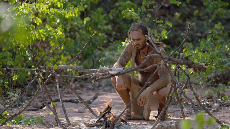 Darrin kneels and stares at the camp fire. – Bild: Warner Bros. Discovery, Inc. or its subsidiaries and affiliates.
