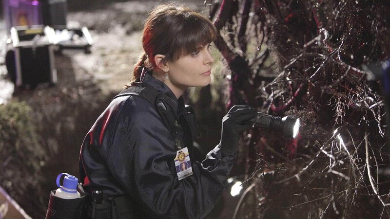 Brennan (Emily Deschanel) untersucht den Fundort einer Leiche, die bereits seit längerer Zeit unter den Wurzeln eines umgefallenen Baumstammes gelegen haben muss. – Bild: TVNOW /​ FOX