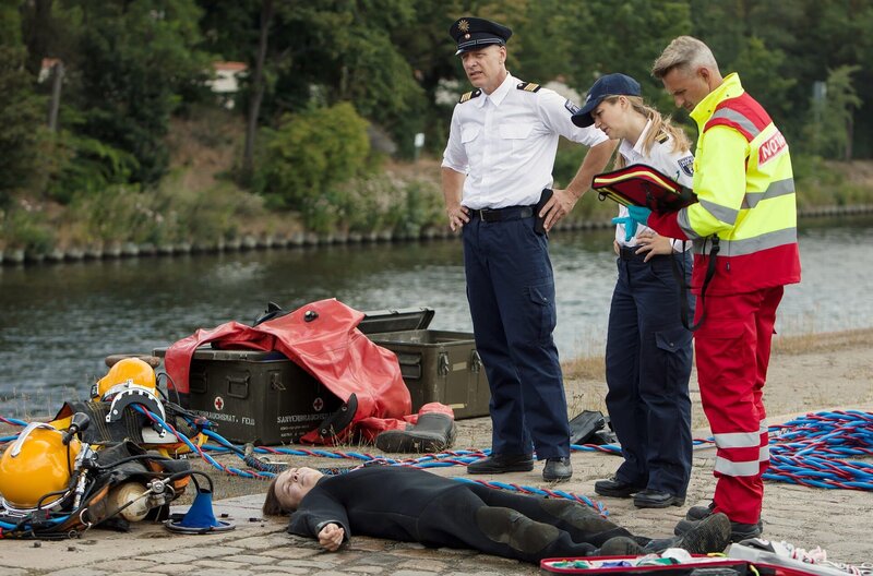 Die erfahrene Industrietaucherin Rosi Neumann ist bei der Instandsetzung der Ufermauer einer Farbenfirma zu Tode gekommen. Paula Sprenger (Sarina Radomski, 2.v.r.) und Wolf Malletzke (Christoph Grunert, 2.v.l.) erhalten erste Informationen vom Notarzt (Komparse, r.). – Bild: ARD/​Daniela Incoronato