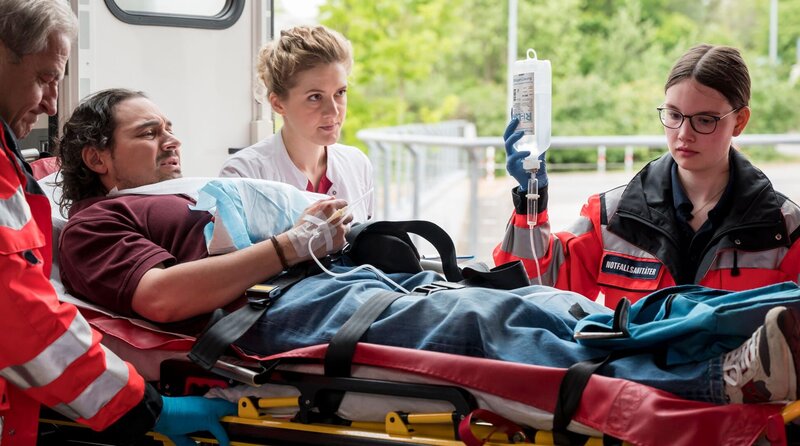 Dr. Elly Winter (Juliane Fisch, M.) nimmt Gideon Dietrich (Johannes Klaußner, 2.v.l. mit Komparse. l.) in der Notaufnahme auf und führt erste Untersuchungen durch (mit Komparsin r.). – Bild: ARD/​Jens-Ulrich Koch