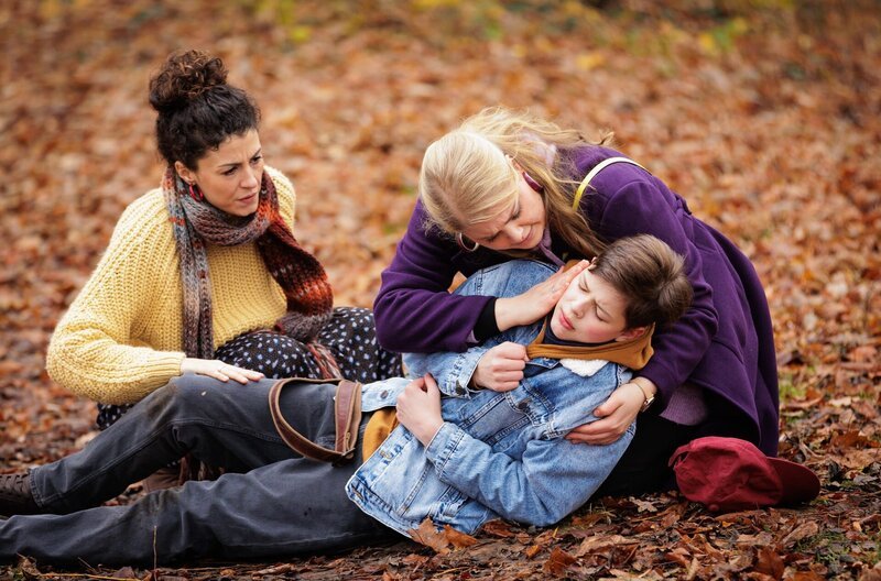 Miriam Schneider (Christina Petersen, re.) und ihre Frau Rieke Machold (Liza Tzschirner, li.) drehen eine Kennenlern-Runde im Park mit ihrem potenziellen Pflegekind Cosmo (Max Bär, liegend). Doch plötzlich bricht der 13-Jährige mit akuter Atemnot zusammen. – Bild: MDR/​Saxonia Media/​Rudolf Wernicke