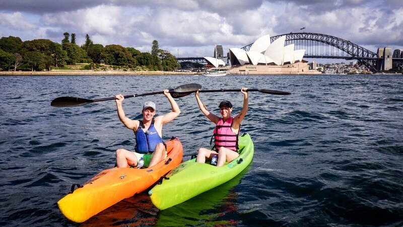 Reiseleiter Moritz und Entertainment Managerin Maika heben, bekleidet mit Schwimmwesten und Kappen, beim Kayak-Fahren vor der Oper und Harbour Bridge in Sydney freudig ihre Paddel in die Luft. – Bild: BR/​Deborah Stöckle