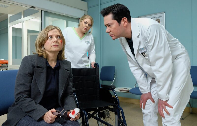 Sylvia Haas (Nina Weniger, li.) kommt mit einer Schnittwunde unter dem Auge, die angeblich von einem heruntergefallenen Bild stammt, in die Sachsenklinik. Dr. Philipp Brentano (Thomas Koch, re.) will die Wunde behandeln und bittet Schwester Miriam (Christina Petersen, mi.) einen Rollstuhl zu bringen. Sylvia scheint sehr wackelig auf den Beinen zu sein. – Bild: BR/​MDR/​Saxonia Media Filmproduktions GmbH/​Sebastian Kiss