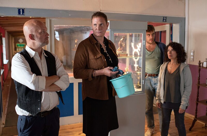 Kegelclubleiter Axel Papst (Björn Jung, li.) missfällt, dass aus seinem Kegelbruder Roman nun Romana (Fabian Harloff, Mi.) geworden ist. Sie hat die Vitrine mit dem „Divers“-Symbol für mehr Toleranz verziert (mit Maria Ketikidou, r. und Enrique Fiß, 2.v.r.). – Bild: ARD/​Thorsten Jander