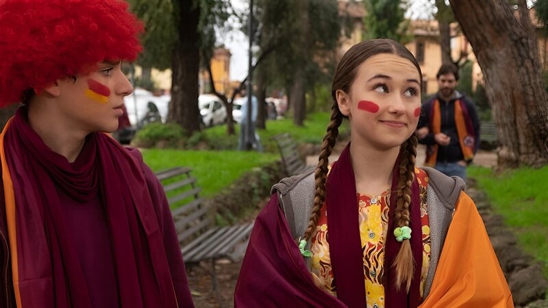 Lucy (Kensington Tallman, r.) und Nico (Christian Monaldi, l.) tragen Schals und Perücken in den Farben ihrer Fußballmannschaft – orange und rot. – Bild: hr/​Red Monk Studios srl/​ HSR S1 (FGM) Inc.