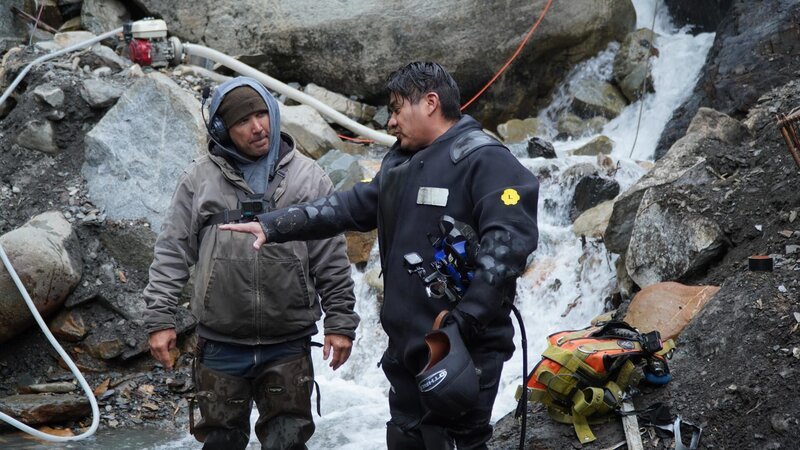 L-R: Carlos Minor conversing with Dustin Hurt next to the water – Bild: Warner Bros. Discovery, Inc. or its subsidiaries and affiliates