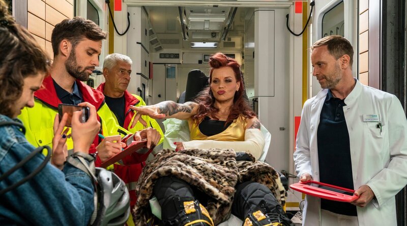 Wie erwartet, wird die Wrestlerin Nikki Klein (Annette Strasser, mi,.) nach ihrem Kampf erneut verletzt in die Sachsenklinik eingeliefert. Dieses Mal ist sehr wahrscheinlich ihr Arm gebrochen. Mit dabei ist wieder ihr Fan Gina Veigel (Amira Demirkiran, li.), die alles aufnimmt und „Bad Nikki“ selbst daraus eine große Show macht. Dr. Kai Hoffmann (Julian Weigend) wird der ganze Trubel schnell zu viel. – Bild: MDR/​Saxonia Media/​Thomas Dietze