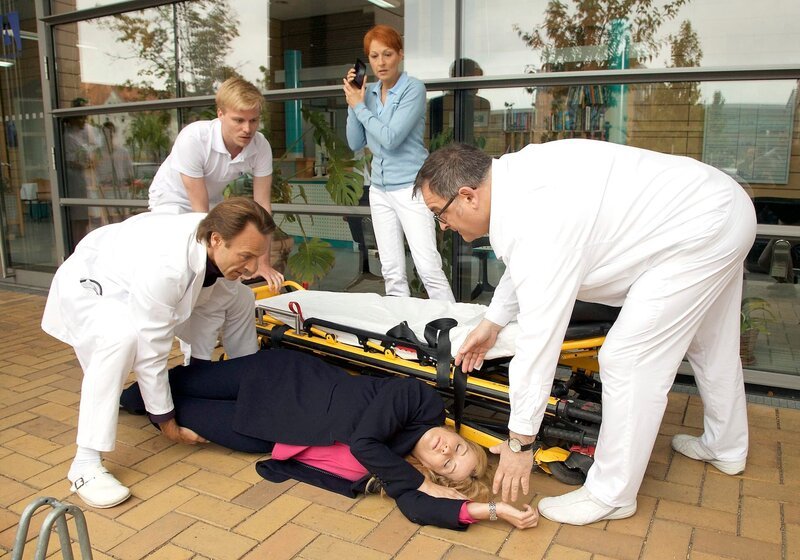Klinikleiterin Sarah Marquardt (Alexa Maria Surholt, liegend) ist vor den Augen von Pia Heilmann (Hendrikje Fitz, Mitte) kollabiert. Dr. Martin Stein (Bernhard Bettermann, vorne links), Hans-Peter Brenner (Michael Trischan, rechts) und ein weiterer Pfleger (Komparse) sind sofort zur Stelle. – Bild: MDR/​Saxonia/​Wernicke