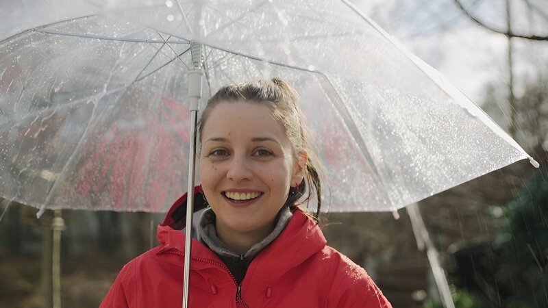 Marina steht bei Regen unter einem Regenschirm. – Bild: BR/​megaherz gmbh/​David Birzer