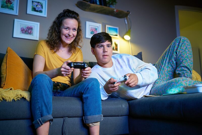 Marie (Tinka Fürst, l.) und Ernst (Diyar Ilhan, r.) sitzen gemeinsam auf dem Sofa und spielen Videospiele. – Bild: ZDF und Guido Engels