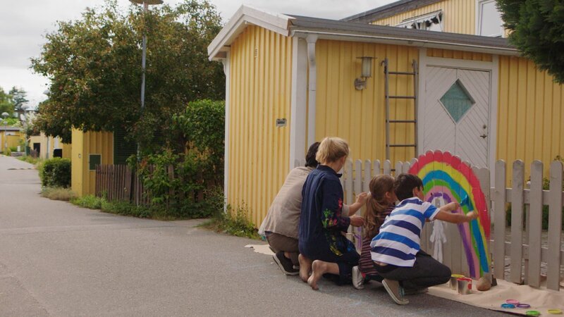 Klara (Paulina Pizarro Swartling) malt mit Mama und Papa einen Regenbogen an den Zaun. – Bild: KiKA/​Erik Vallsten