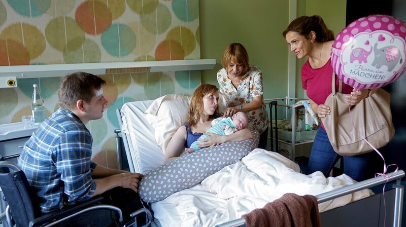 Vera (Rebecca Immanuel, re.) und Bärbel Kuhn (Gitta Schweighöfer) gratulieren den frisch gebackenen Eltern Simon (Enno Hesse, li.) und Lena (Christin Nichols). – Bild: ARD Degeto/​Hans-Joachim Pfeiffer