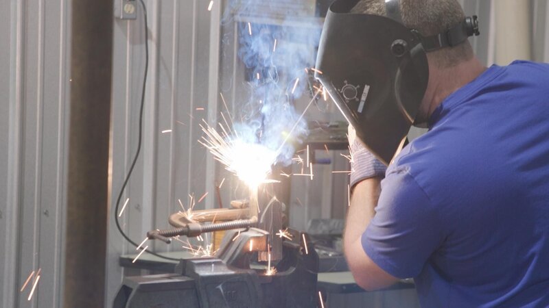 Ein Mann, der einen Schutzhelm auf dem Gesicht trägt, schmiedet Eisen – Bild: A&E Networks, LLC.