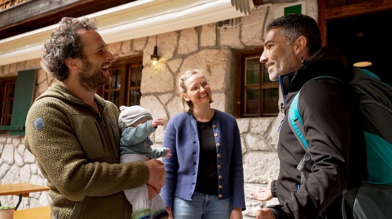 Die Reintalangerhütte im Wettersteingebirge: Moderator Marcus Fahn (rechts) auf der Terasse mit den Hüttenwirtsleuten Andy und Julia Kechle und ihrem Sohn. – Bild: BR/​Watch Lola GmbH/​Matthias Schwinn
