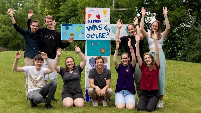 Checker Tobi mit vielen Ministranten und Ministrantinnen vor einem selbstgebastelten Kreuz. – Bild: BR/​megaherz gmbh/​Sarah Müller