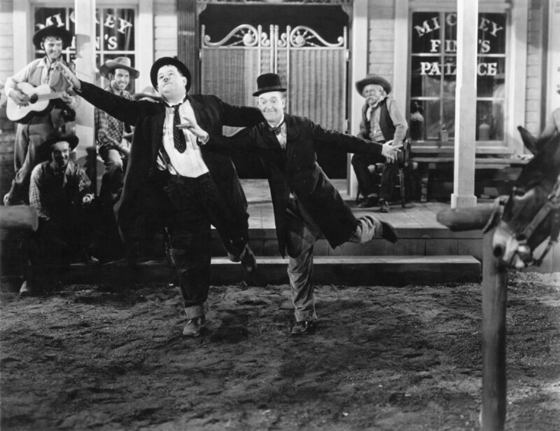 Als Stan (Stan Laurel – im Vordergrund rechts) und Ollie (Oliver Hardy – im Vordergrund links) in Brushwood Gulsh ankommen, machen einige Cowboys (Darsteller im Hintergrund unbekannt) Musik vor dem Saloon. Das animiert beide, ein Tänzchen einzulegen. – Bild: ZDF und Stax Graves