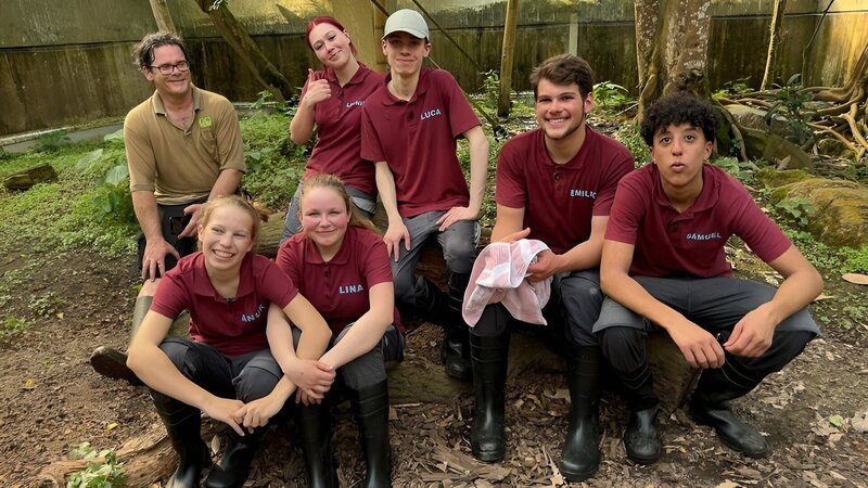 Michael Ernst, Anuk, Lina, Leonie, Luca, Emilio und Samuel (v. l. n. r.) sitzen auf Baumstamm nach der Otterbeckensäuberung im Zoo Leipzig. – Bild: MDR/​Cine Impuls Leipzig GmbH