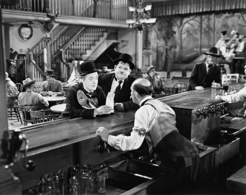 Im Saloon erkundigen sich Stan (Stan Laurel – links an der Theke) und Ollie (Oliver Hardy – rechts an der Theke) beim Besitzer (James Finlayson – mit dem Rücken zu Kamera) nach Mary Roberts, der sie ihre Erbschaft überbringen sollen. Obwohl es ein Geheimnis sein soll, worum es sich dabei handelt, plaudert es Stan sofort aus. – Bild: ZDF und Stax Graves