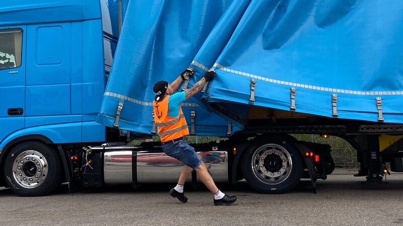 Der Fahrer richtet die Plane auf dem Lkw aus. – Bild: Warner Bros. Discovery