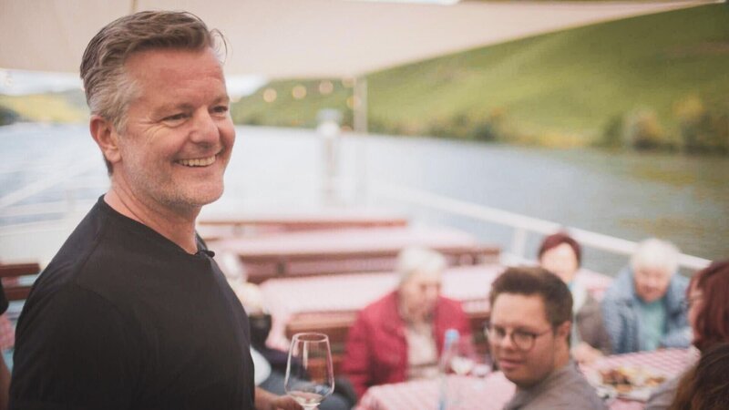 André Dietz lacht auf Schiff im Hintergrund Pfleger:innen und Senor:innen – Bild: RTL /​ Folge 01_004
