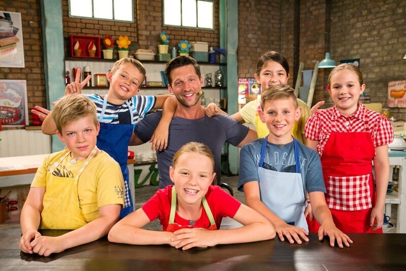 Fernsehkoch und Moderator Alexander Kumptner zeigt den Koch-Kids verschiedene Gerichte mit Bananen. – Bild: ZDF/​Rene Rothkopf