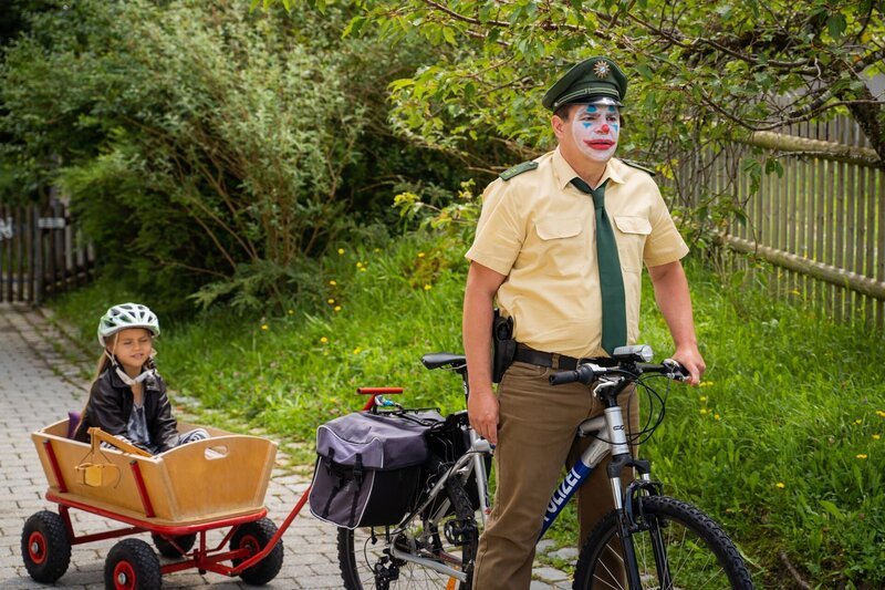 Riedl (Paul Sedlmeir) bemerkt nicht, dass er aussieht wie ein Clown und macht sich mit Lina (Carla Graßl) im Bollerwagen auf den Weg zum Revier. – Bild: Leonine /​ Jenrick Mielke