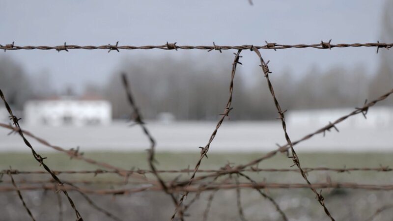 Zaun mit Sicht auf Baracke. – Bild: BR/​Tellux-Film GmbH/​Tobias Tempel