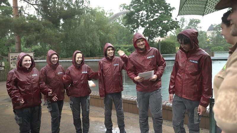 Lina, Leonie, Anuk, Luca, Emilio, Samuel, Meriam Pietsch und Christoph Urban stehen mit Teilnahmeurkunden im Regen bei Verabschiedung vom Praktikum vor dem Seelöwengehege des Zoo Leipzigs. – Bild: MDR/​Cine Impuls Leipzig GmbH