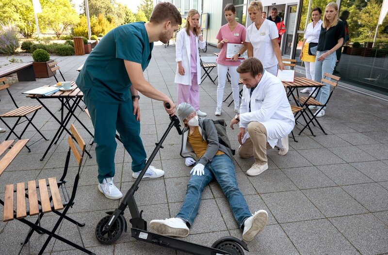 Dr. Marc Lindner (Christian Beermann, v.r.) und Mikko Rantala (Luan Gummich, v.l.) sitzen in der Cafeteria, als Joshy Binder (Laurids Schürmann, v.M.) mit seinem Roller stürzt und sich verletzt (mit Kompars:innen, h.). – Bild: ARD/​Jens-Ulrich Koch