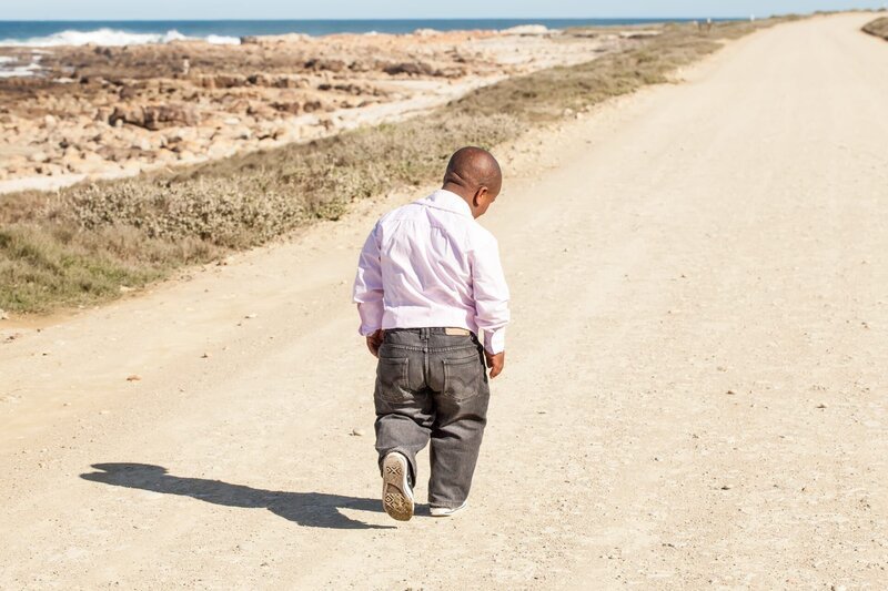 dwarfish african man walking down a dirt road along the coast with his head bent down – Bild: Nolte Lourens