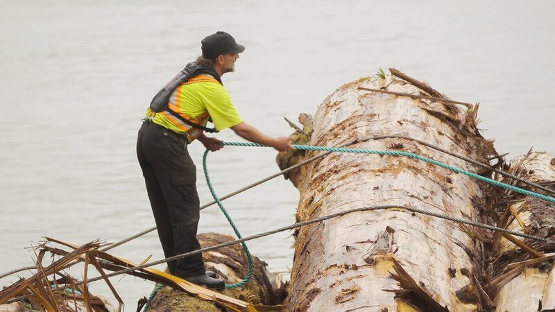 Dave Lornie stands on a floating log bundle preparing to lash a tow rope to the bundle. – Bild: Warner Bros. Discovery, Inc. or its subsidiaries and affiliates. All rights reserved.