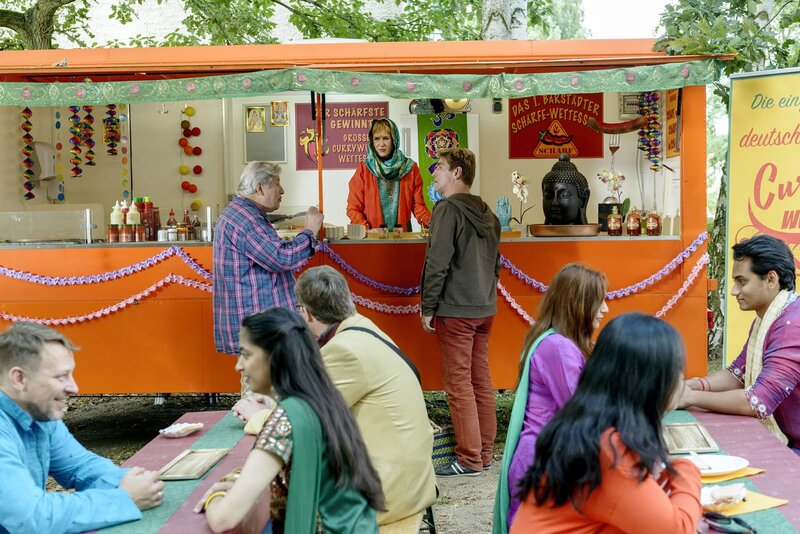 Am indischen Currywurst-Stand von Indira (Beatrice Richter, hinten Mitte) treffen sich die beiden Streithähne Fritz Fuchs (hinten, r.) und Nachbar Paschulke (hinten, l.) fürs Schärfewettessen. – Bild: ZDF/​Antje Dittmann